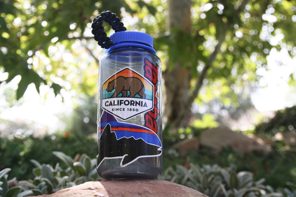 A clear water bottle on a rock carries the California State Bear sticker beside a trout decal.
