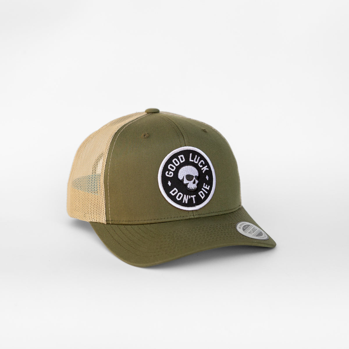 The Good Luck Don't Die trucker hat sits in olive green with a skull patch stitched above its tan mesh back.