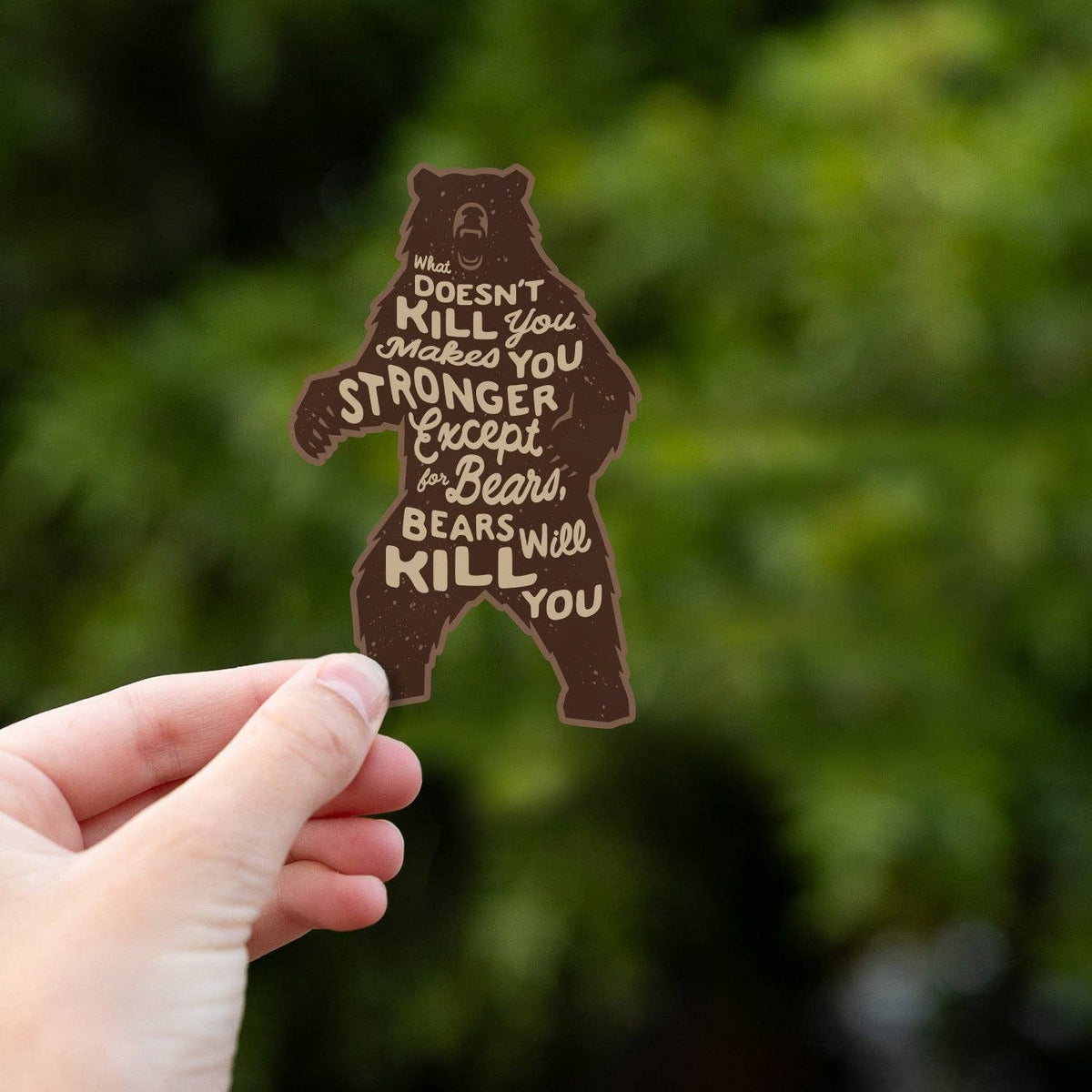 A hand holds the Bears Will Kill You sticker up against blurred green leaves.