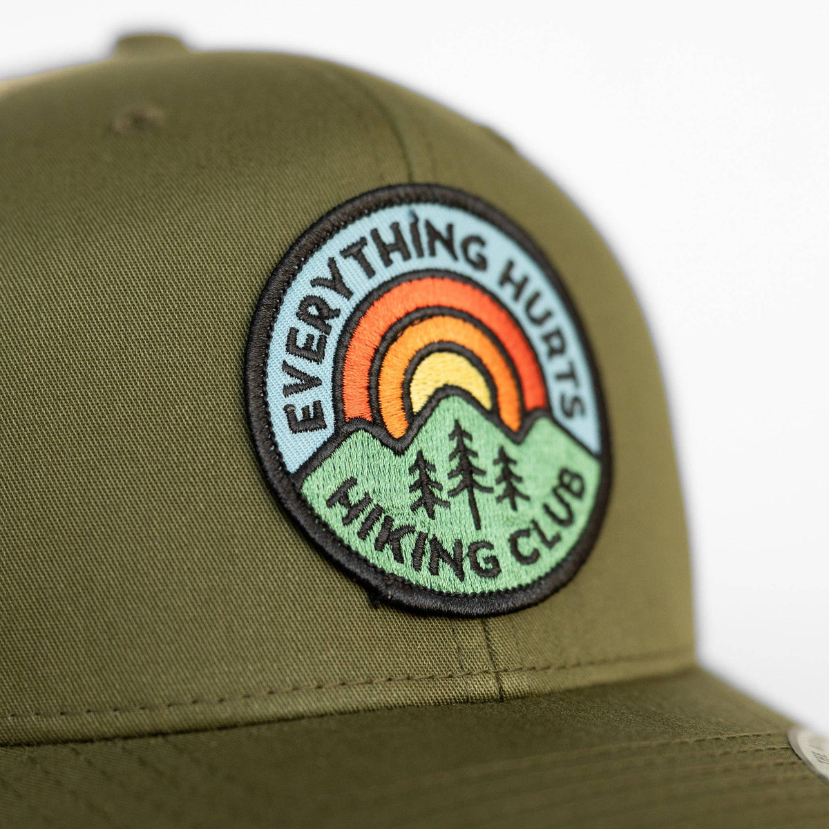 The patch reads Everything Hurts Hiking Club above green mountains on the olive crown of the trucker hat.