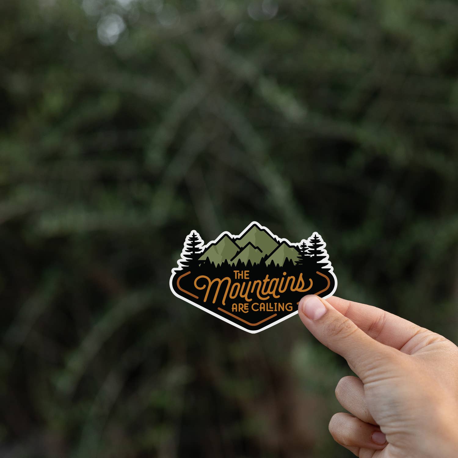 A fingertip balances The Mountains Are Calling sticker against blurred dark evergreen branches.