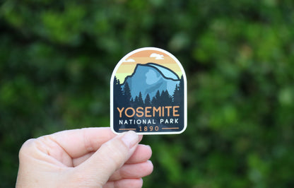Fingers pinch the lower edge of the Yosemite National Park sticker, greenery soft behind it.