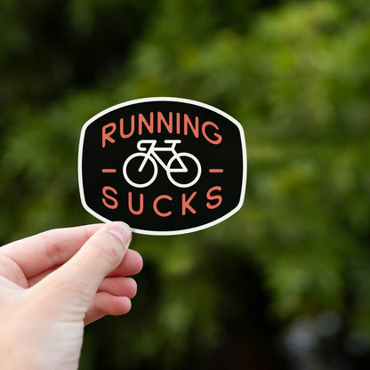 Fingers grip the lower edge of the Running Sucks sticker in front of green summer foliage.