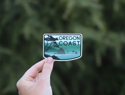 Fingers pinch the Oregon Coast sticker at arm's length, small against a wall of dark evergreen branches.