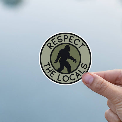 Against calm grey water, fingers hold the Bigfoot Respect The Locals sticker steady in the light.