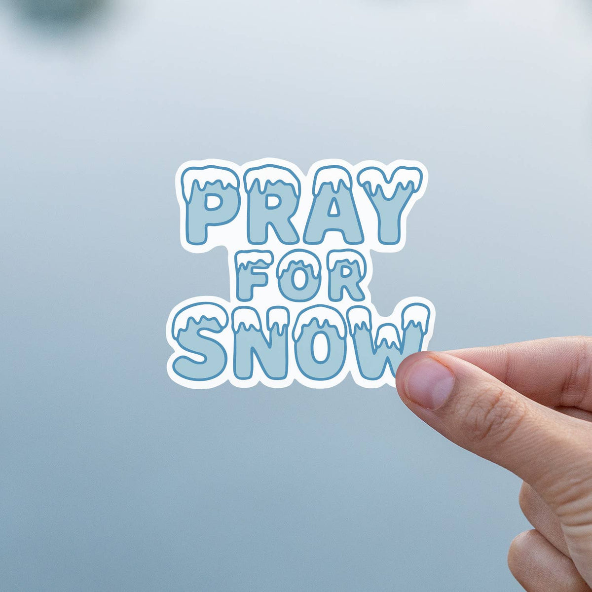 Fingers hold the Pray For Snow sticker against a soft grey background indoors.