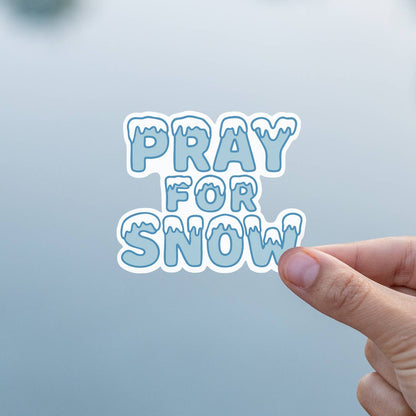 Fingers hold the Pray For Snow sticker against a soft grey background indoors.
