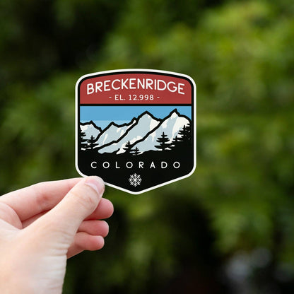 Fingers grip the lower edge of the Breckenridge Colorado sticker with leafy trees blurred behind.