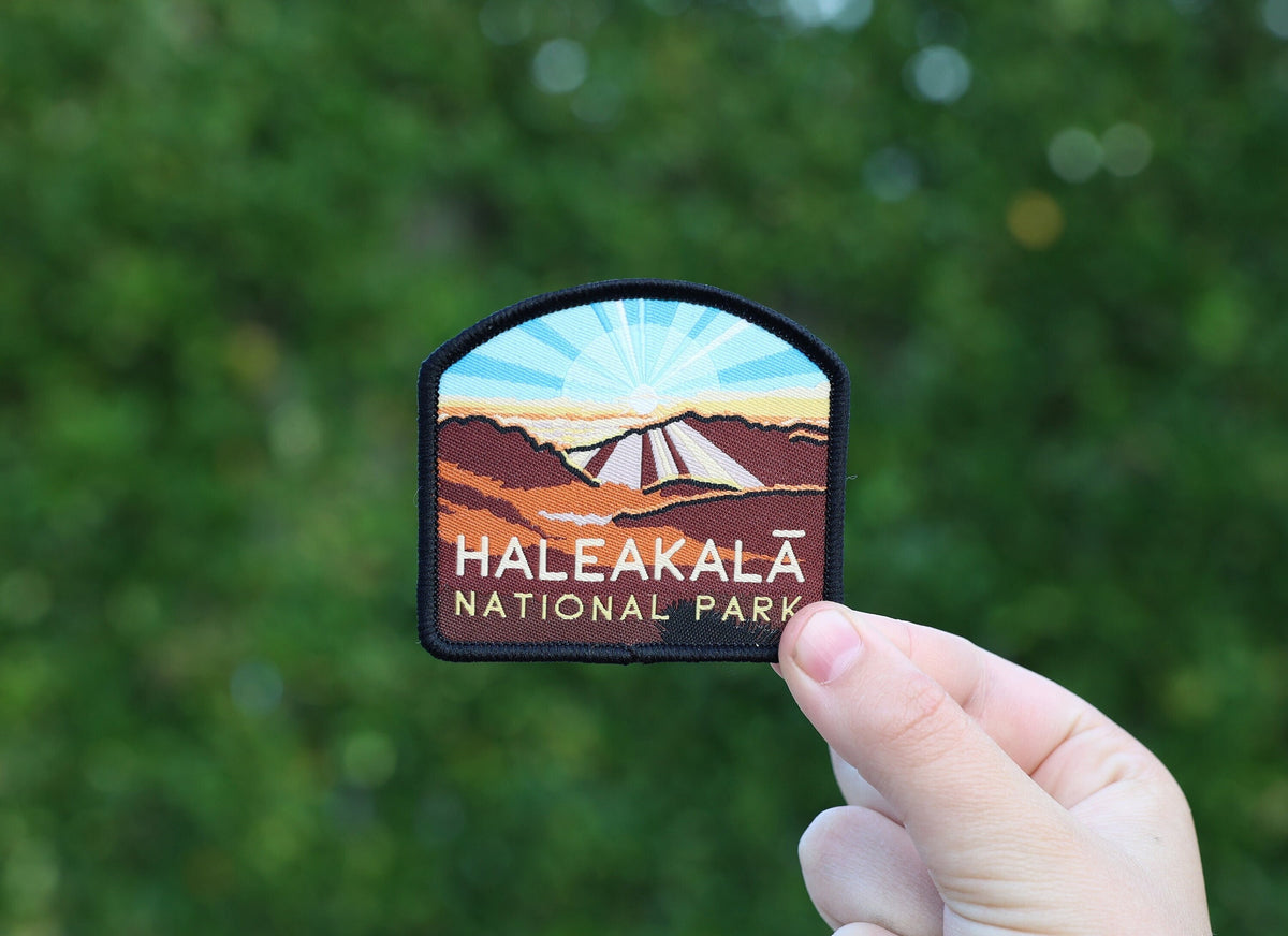 Fingers hold the Haleakala National Park patch up against a blurred green wall of leaves.