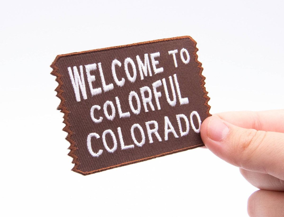 Fingers hold the Welcome to Colorful Colorado patch at an angle, showing its small size against a pale background.