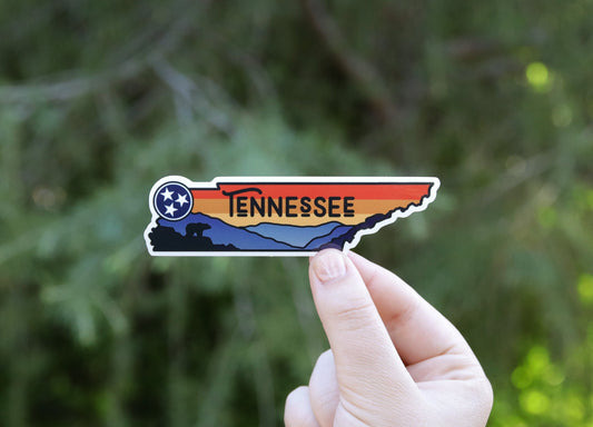 Held further back against evergreen branches, the Tennessee State sticker shows its tristar and blue mountains.