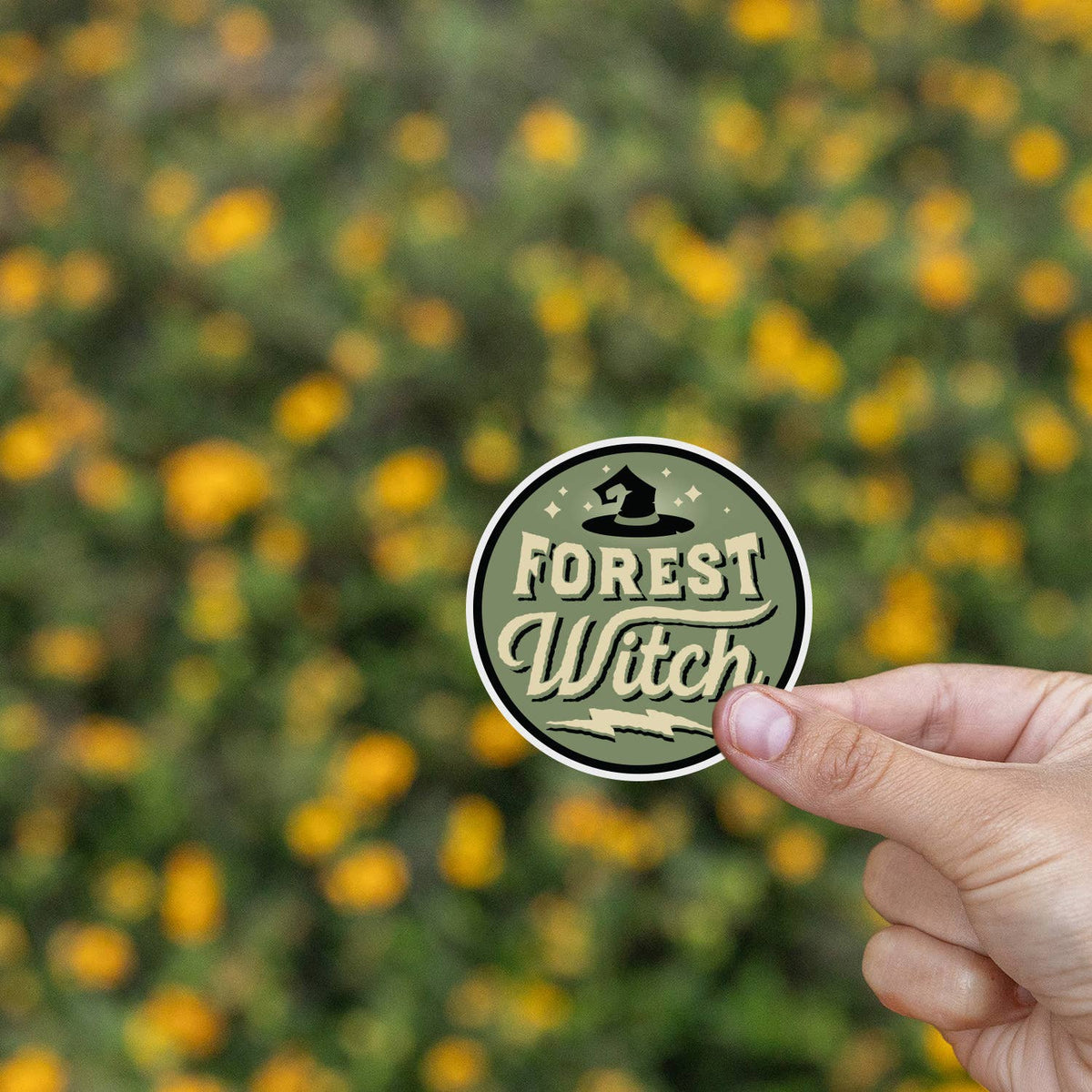 A field of yellow wildflowers blurs behind the Forest Witch sticker held at arm's length.