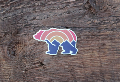 Pressed flat on weathered brown wood, the Adventure Grizzly Bear sticker shows its pink and blue tones.
