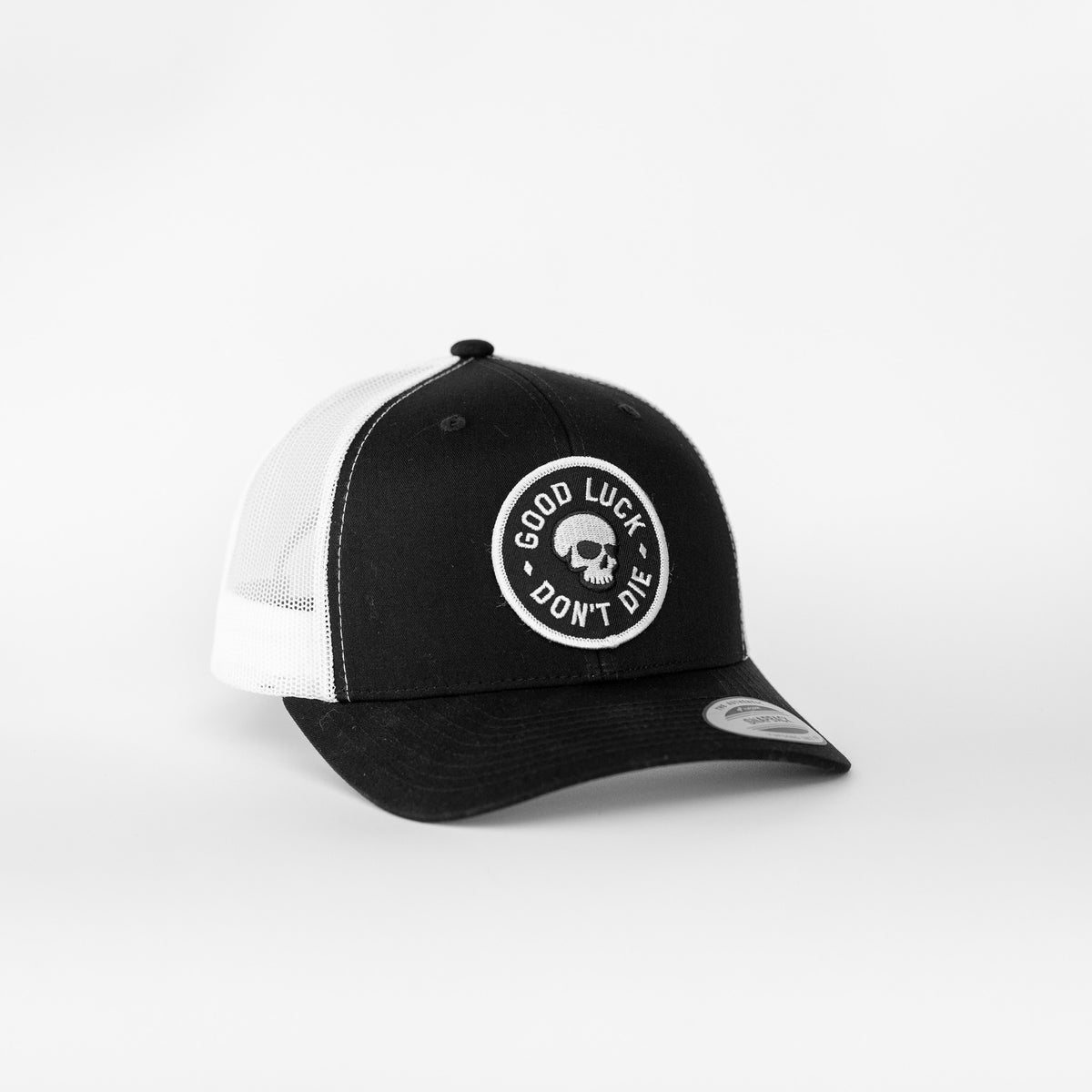 Black and white panels split the Good Luck Don't Die trucker hat, the skull patch centered on the dark front.