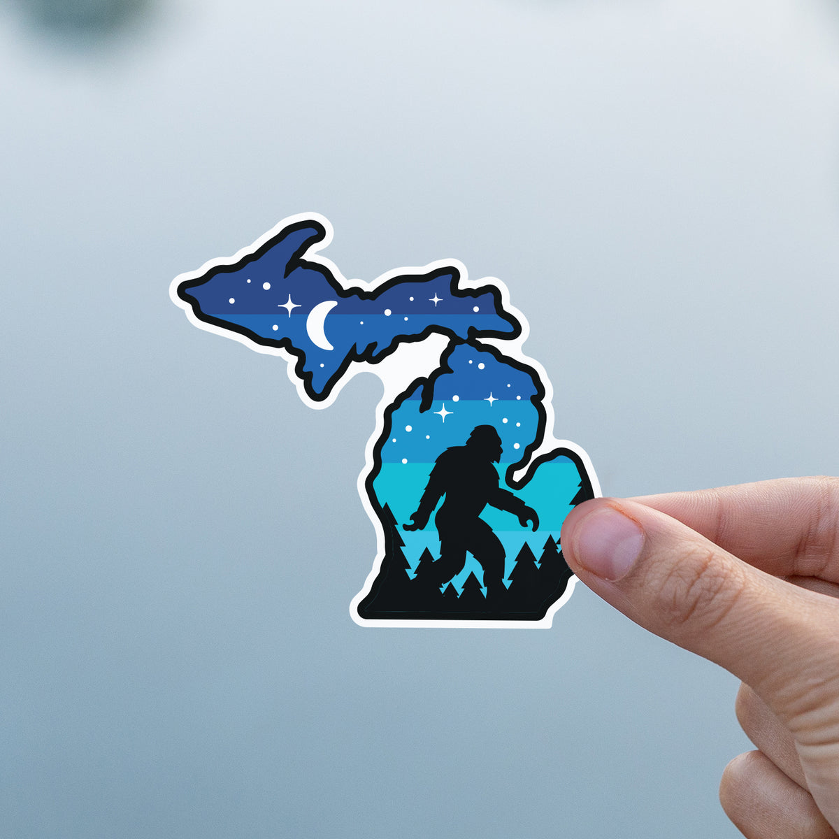 A thumb and finger present the Michigan Sasquatch sticker against a flat grey body of water.