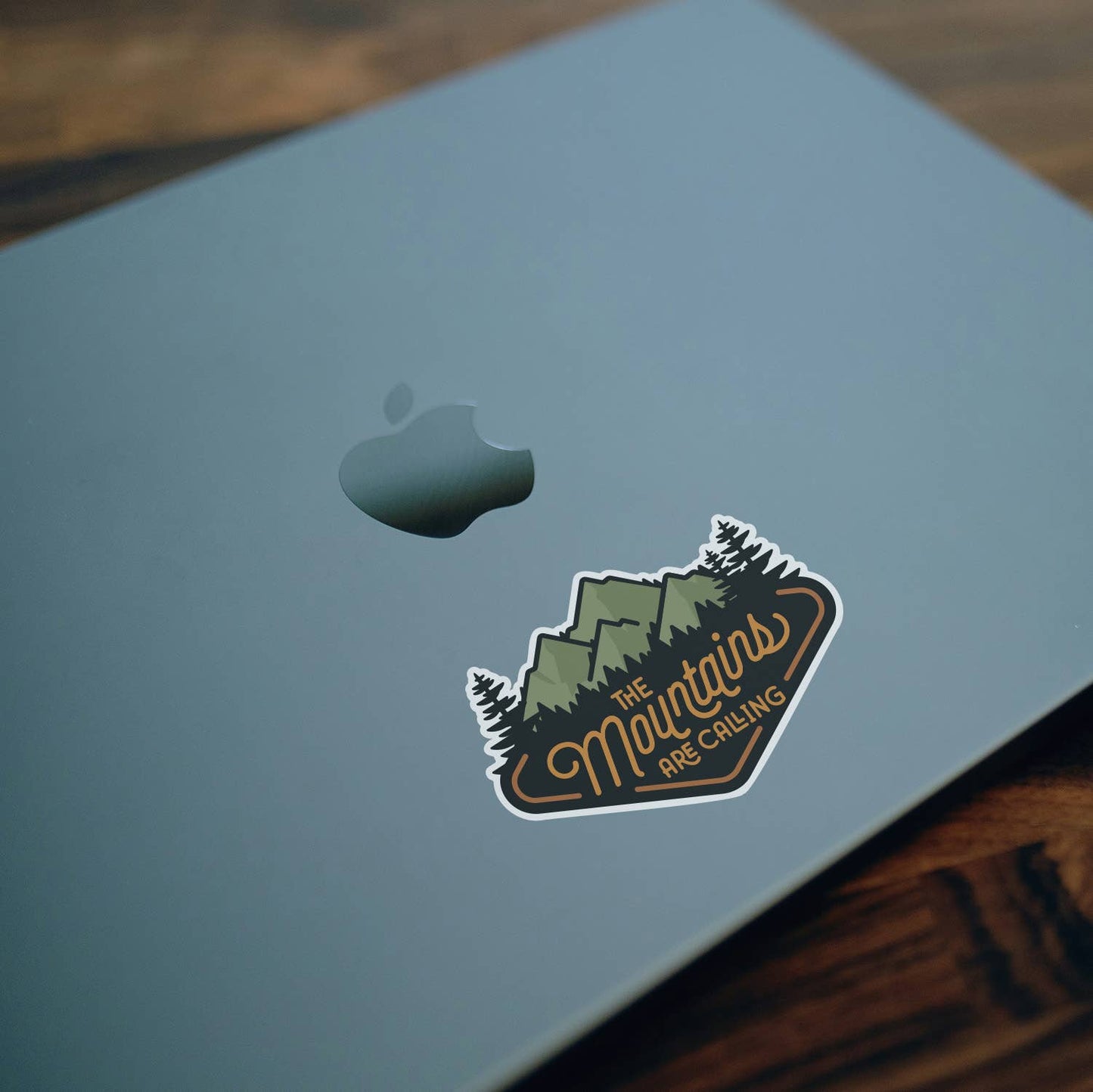 Stuck near the logo on a pale blue laptop lid, The Mountains Are Calling sticker rests over dark wood.