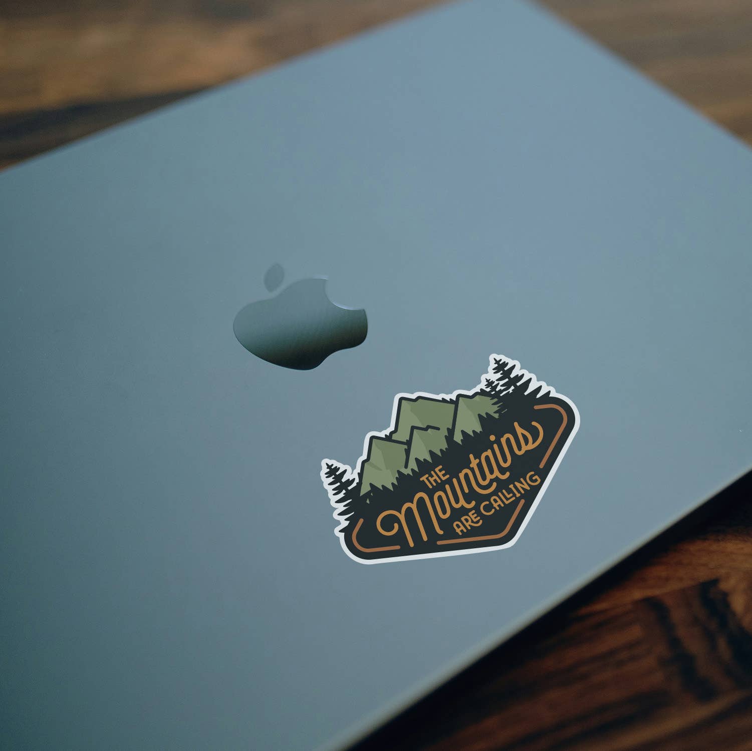 Stuck near the logo on a pale blue laptop lid, The Mountains Are Calling sticker rests over dark wood.
