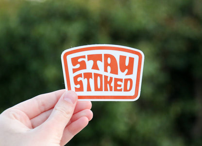Fingers grip the Stay Stoked sticker close to the lens, with a hedge blurred behind it.