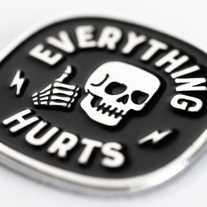 A close macro view shows the Everything Hurts enamel pin, its grinning skull and bony thumbs up catching the light.