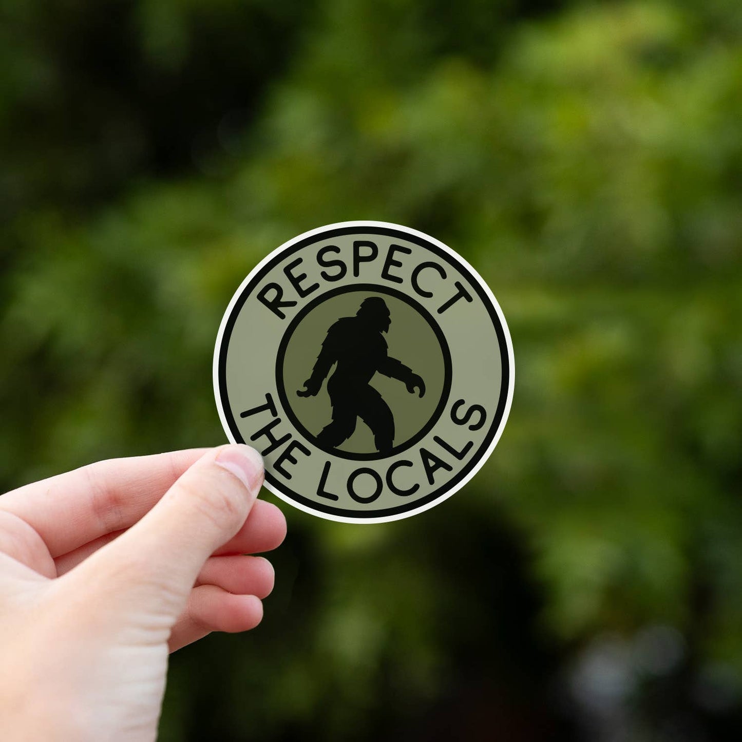 Held close to the lens, the Bigfoot Respect The Locals sticker fills the frame against sunlit green leaves.