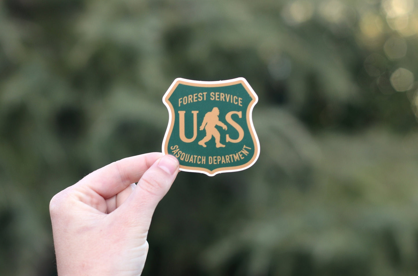 Pale pine needles fall away behind the US Sasquatch Department sticker held in a raised hand.