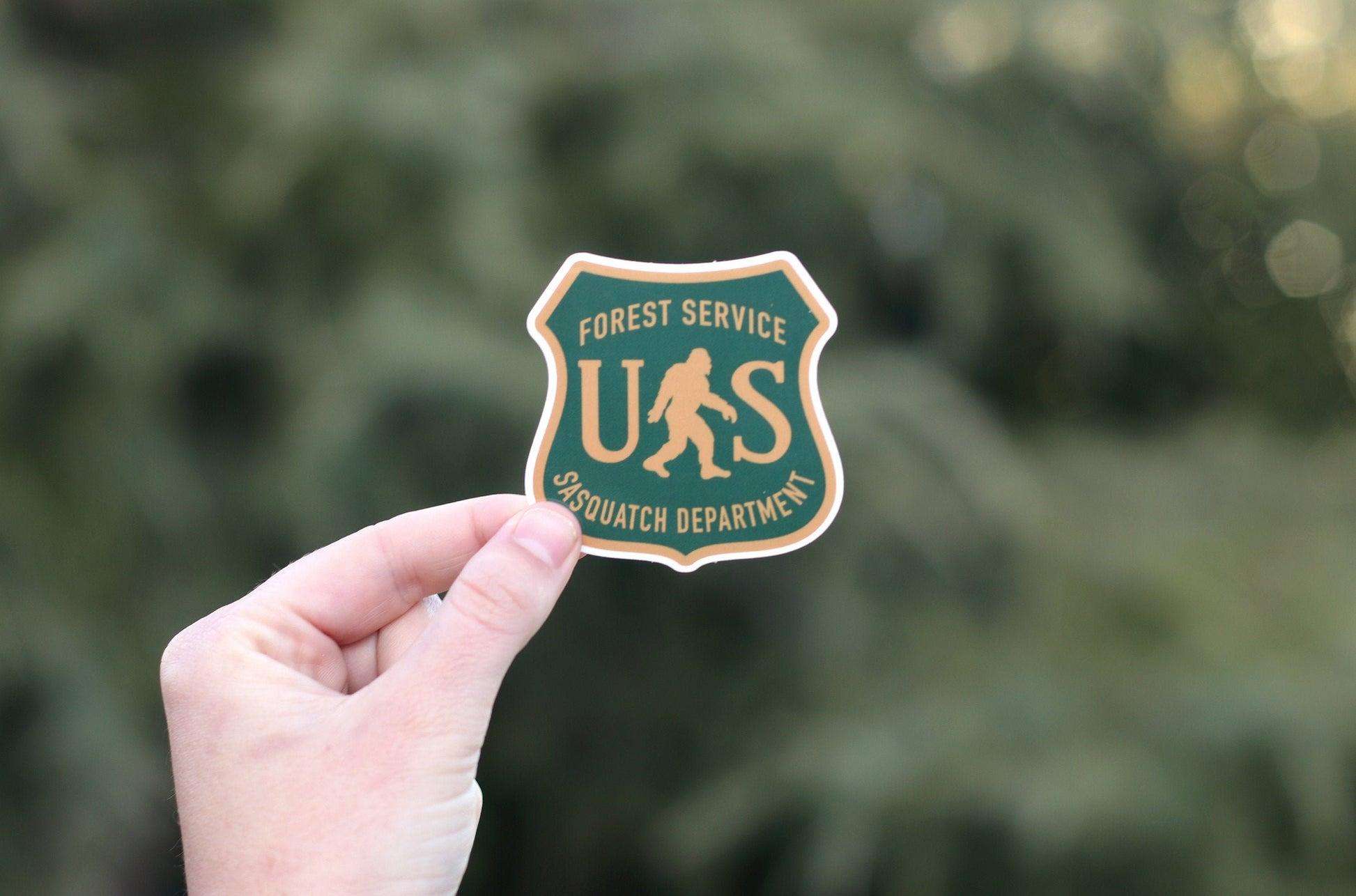Pale pine needles fall away behind the US Sasquatch Department sticker held in a raised hand.