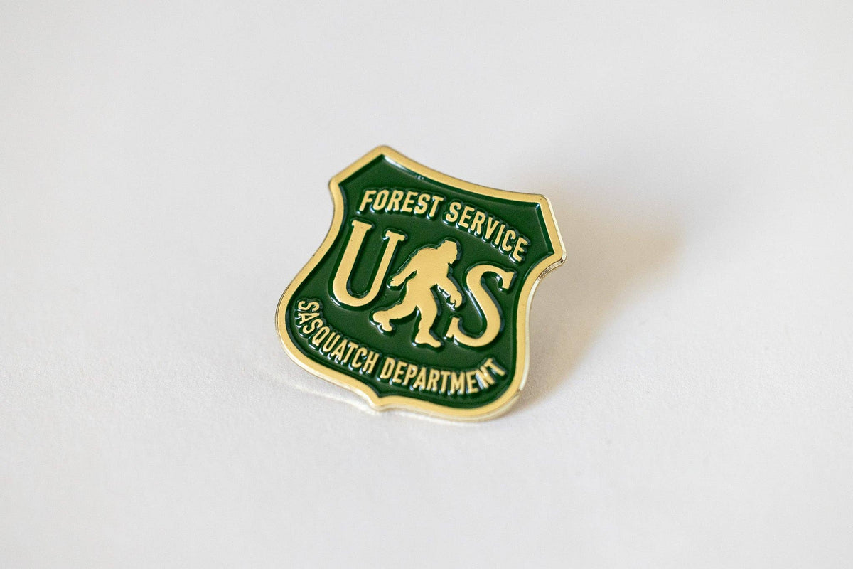 Angled on a plain white surface, the US Sasquatch Department pin catches light along its gold shield edge.