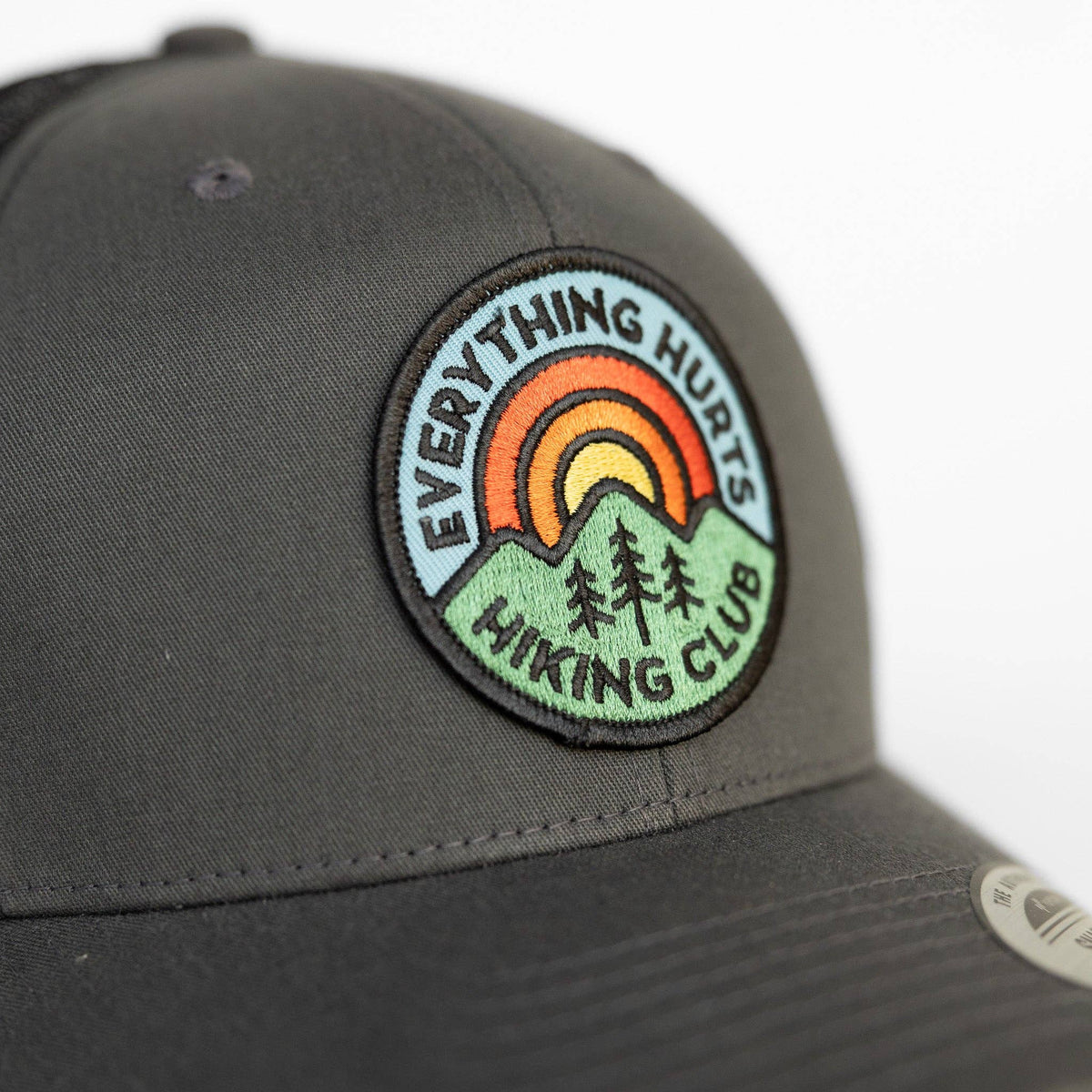 Close on the round sunrise and pine patch stitched to the grey Everything Hurts Hiking Club trucker hat.