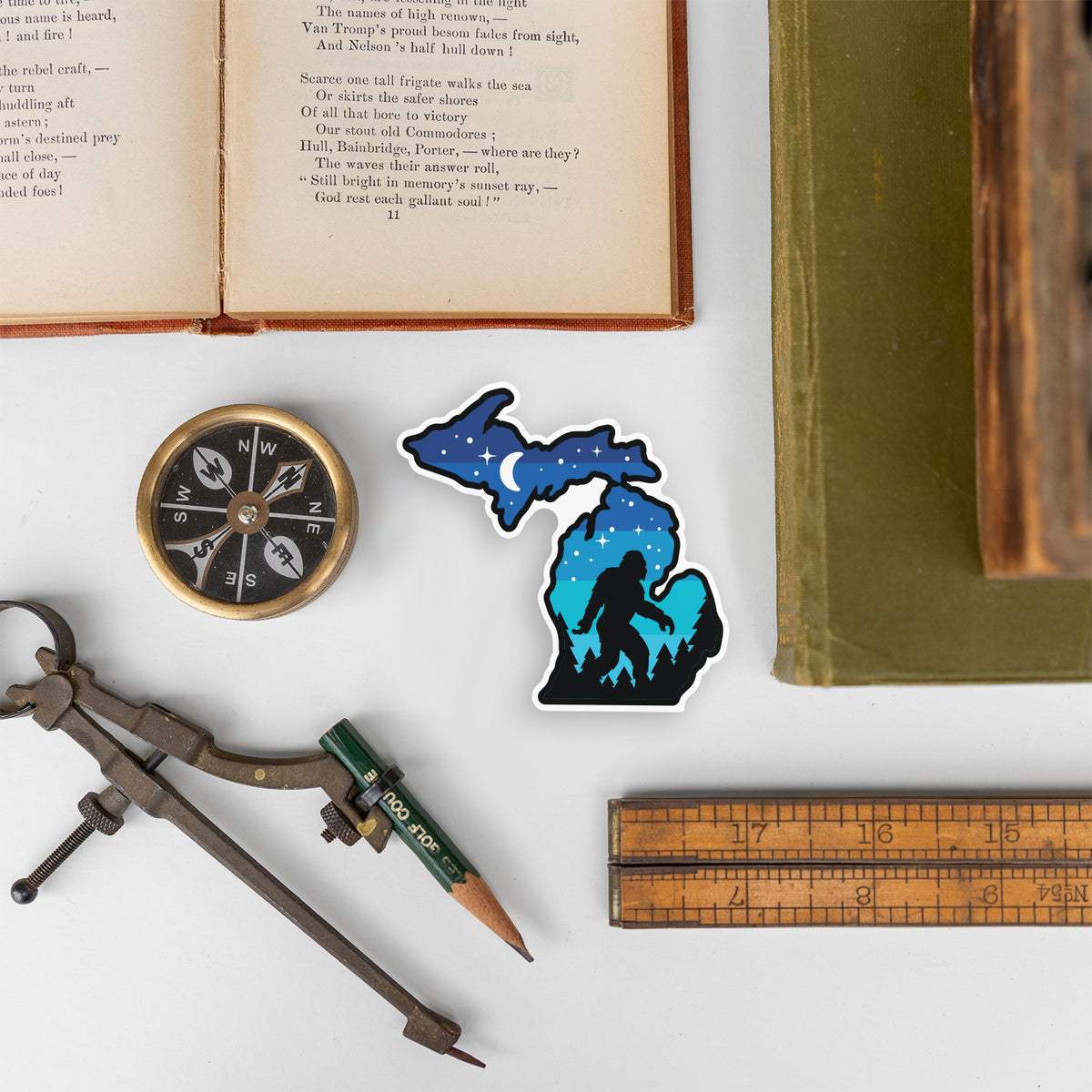 The Michigan Sasquatch sticker rests among an open book, compass, ruler, and drafting tools on a white surface.