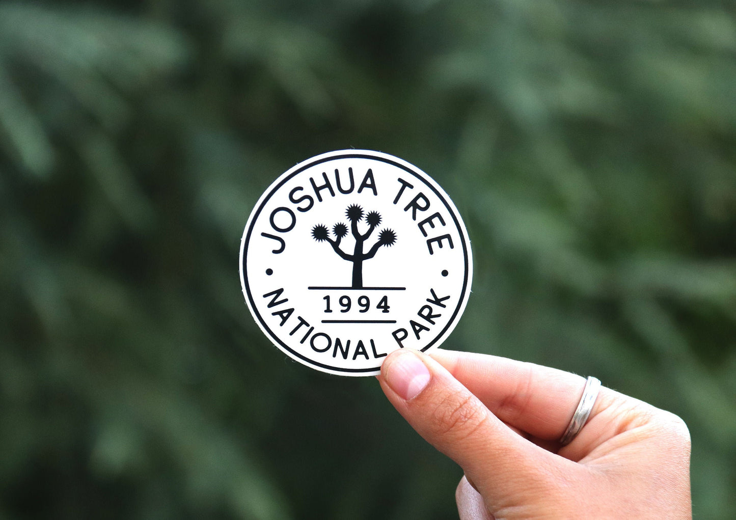 A hand holds the round Joshua Tree National Park sticker, black and white with a 1994 date, against blurred green foliage.