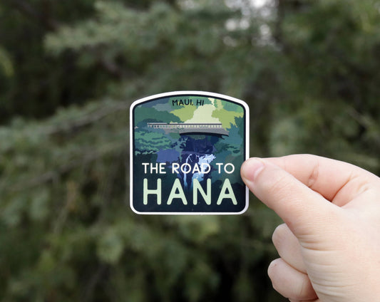 A hand holds the Road to Hana sticker against blurred evergreen branches, its bridge and waterfall art in view.