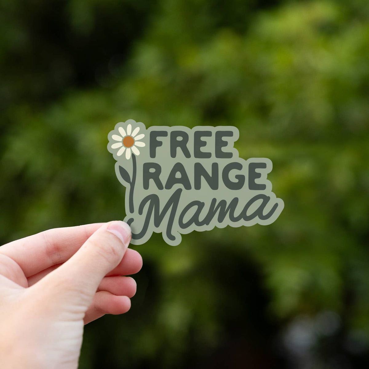 Close and centered, the Free Range Mama sticker rests in a hand with green leaves blurred behind it.