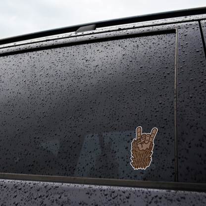 Rain beads across a black car window where the Bigfoot Rocks sticker is applied near the glass edge.