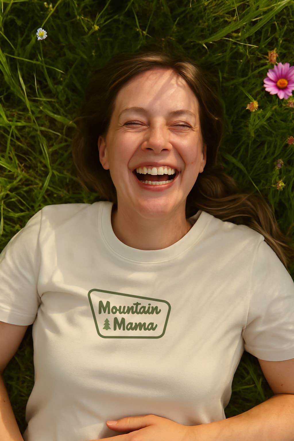 Laughing in the grass among daisies, a woman wears the Mountain Mama T shirt in cream cotton.