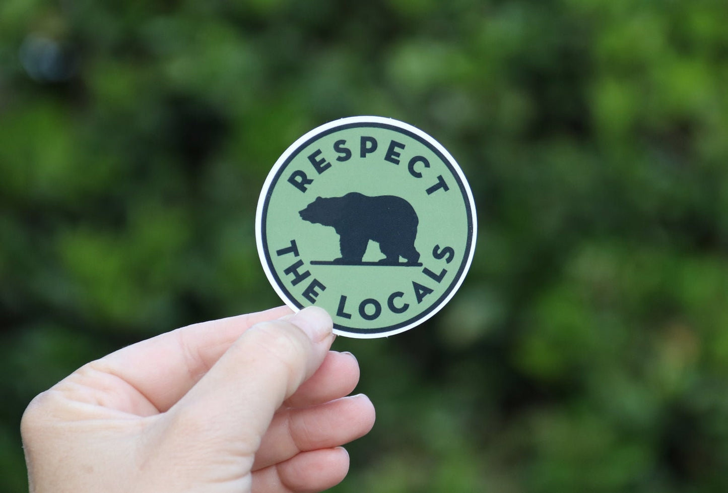 A hand holds the green Respect The Locals sticker with its black bear silhouette against leafy shade.