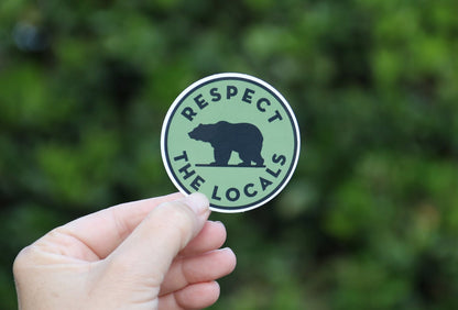 A hand holds the green Respect The Locals sticker with its black bear silhouette against leafy shade.