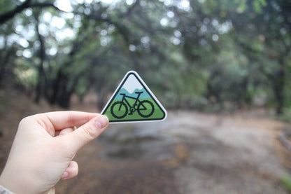 On a damp dirt trail under oak branches, a hand holds the triangular MTB sticker.