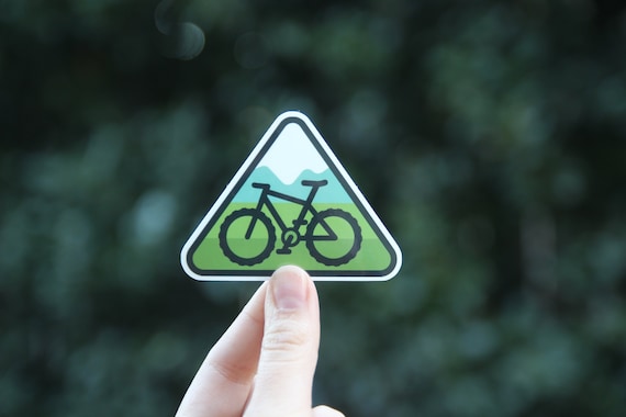 Fingertips lift the triangular mountain bike trail sticker close to the camera against dark leaves.