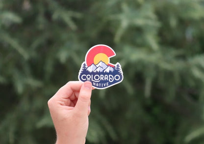 A hand holds up the Colorado Native sticker with its red C and snowy peaks against a blurred evergreen.