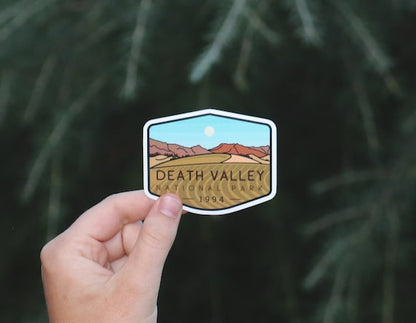 A hand holds the Death Valley National Park sticker with its dunes and red hills against blurred pine boughs.