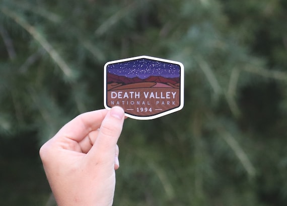 A hand holds the Death Valley National Park at night sticker against blurred green pine branches.