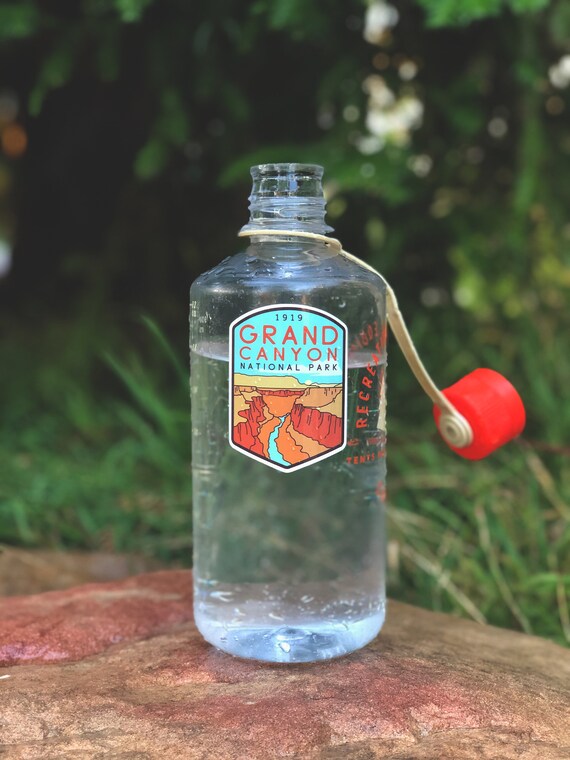A clear water bottle on a rock wears the Grand Canyon National Park sticker, red cap resting against the grass.