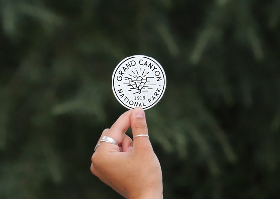 A ringed hand holds the black and white Grand Canyon National Park sticker against blurred evergreen branches.