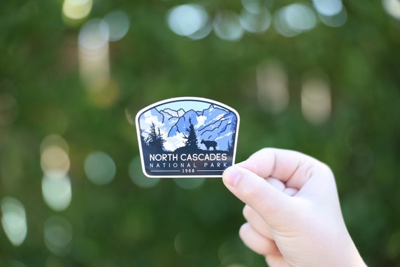 A hand holds the North Cascades National Park sticker against blurred green foliage, its blue peaks and pines in view.