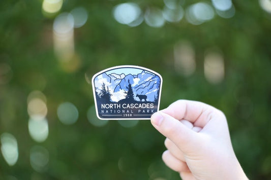 A hand holds the North Cascades National Park sticker against blurred green foliage, its blue peaks and pines in view.