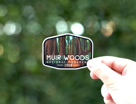 A hand holds the Muir Woods National Monument sticker with its redwood trunks against blurred green foliage.