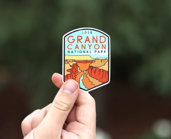 Pinched low between finger and thumb, the Grand Canyon National Park sticker tilts toward a dim woodland backdrop.