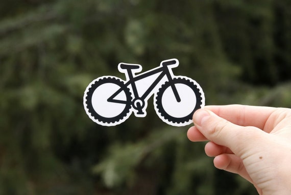 The black and white mountain bike shaped sticker is pinched in front of a dense evergreen.