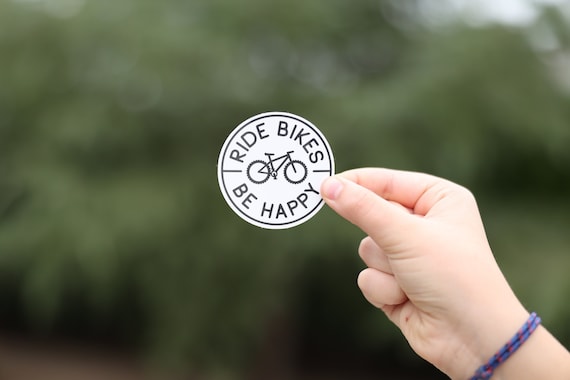 A hand with a braided bracelet holds up the Ride Bikes Be Happy sticker outdoors.
