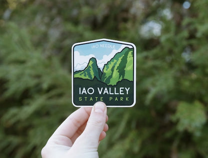 A hand holds the Iao Valley Sticker against blurred green foliage, its Iao Needle peaks in view.